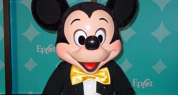 Mickey Mouse in a tuxedo and yellow bow tie greets guests against an Epcot backdrop, ready for your magical Disney World adventure.