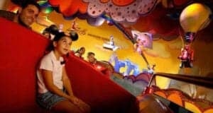 A young girl wearing Mickey Mouse ears and a man ride in a vibrant, whimsical amusement park attraction featuring colorful decorations, playful characters, hot air balloons, and catchy Disney songs, creating a joyful and animated atmosphere.