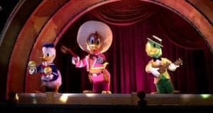 Three colorful Disney animatronic characters—Donald Duck with maracas, Panchito in a sombrero, and a Jose Carioca with a guitar—stand on stage, ready to perform catchy Disney songs in front of red curtains under an arched set.