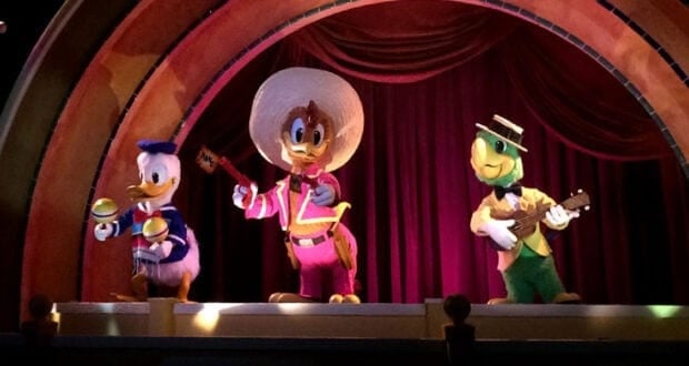 Three colorful Disney animatronic characters—Donald Duck with maracas, Panchito in a sombrero, and a Jose Carioca with a guitar—stand on stage, ready to perform catchy Disney songs in front of red curtains under an arched set.