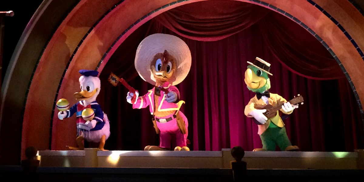 Three colorful Disney animatronic characters—Donald Duck with maracas, Panchito in a sombrero, and a Jose Carioca with a guitar—stand on stage, ready to perform catchy Disney songs in front of red curtains under an arched set.
