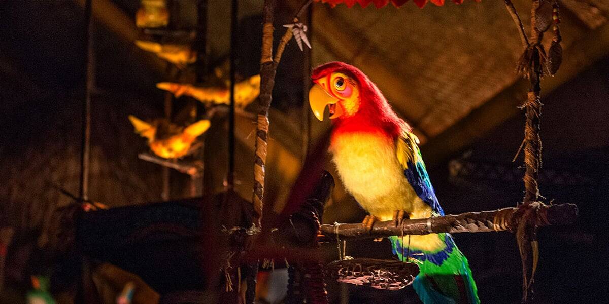 A colorful animatronic parrot with red, yellow, blue, and green feathers sits on a perch in a dimly lit, tropical-themed setting as catchy Disney songs play. Other bird figures are visible in the background.