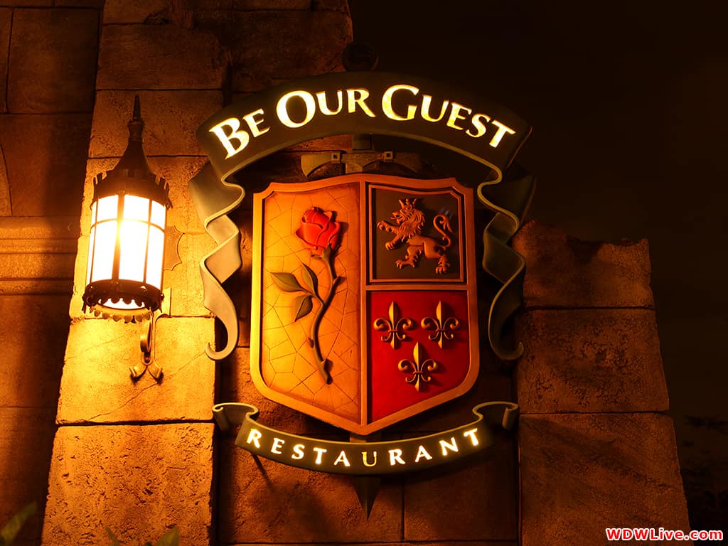 The "Be Our Guest Restaurant" sign glows under a rose, lion, and fleur-de-lis crest at Magic Kingdom, welcoming Disney fans.