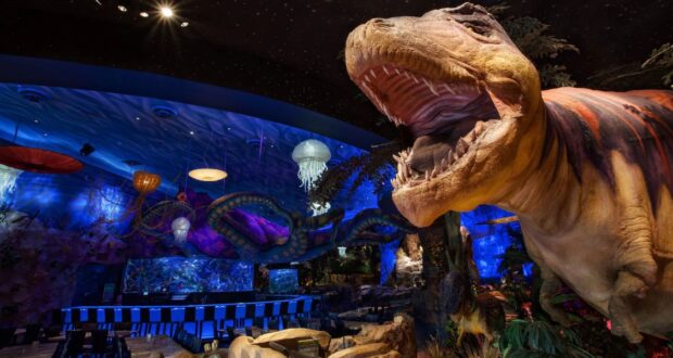 A vibrant themed restaurant interior featuring a large T-Rex model with its mouth open. The atmosphere is enhanced by dim lighting, glowing jellyfish decorations on the ceiling, and a long bar with stools in the background.