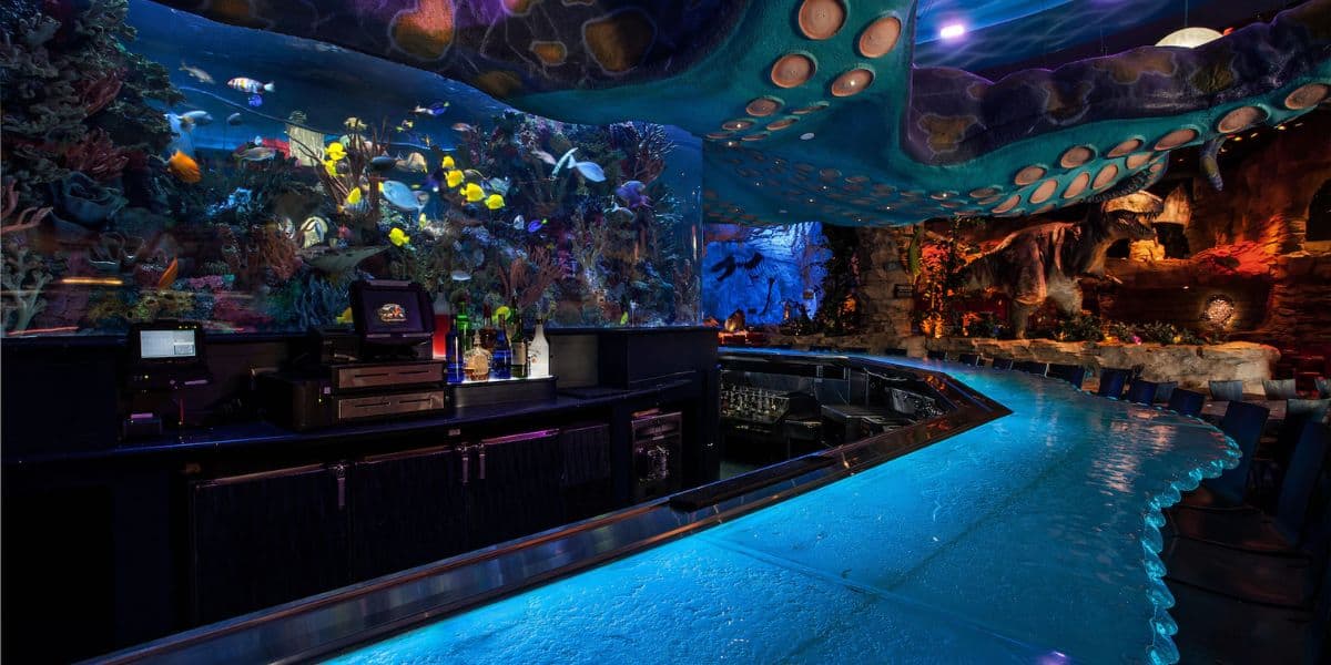 Aquatic-themed bar with a glowing blue countertop, surrounded by stools. An expansive aquarium serves as the backdrop, filled with vibrant fish and coral. Dim lighting and rustic decor enhance the underwater ambiance.