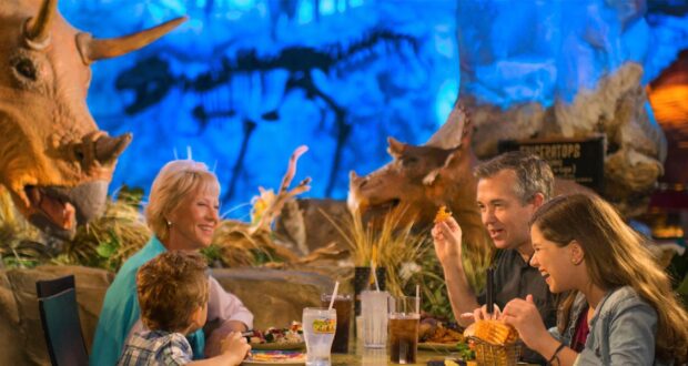 A family of four enjoys a meal at a themed restaurant with dinosaur decor. Two adult figures and two children are seated around a table, smiling and interacting. A triceratops statue is visible in the background against a blue-lit setting.
