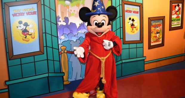Mickey Mouse in his red robe and blue wizard hat greets guests with magical energy amid vibrant Disney World backdrops.