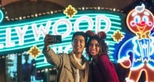 A smiling young couple in festive outfits takes a selfie at night in front of a brightly lit Hollywood sign and a large glowing gingerbread man decoration.