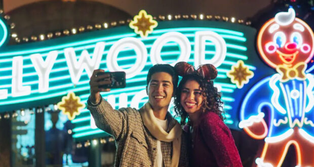 A smiling young couple in festive outfits takes a selfie at night in front of a brightly lit Hollywood sign and a large glowing gingerbread man decoration.