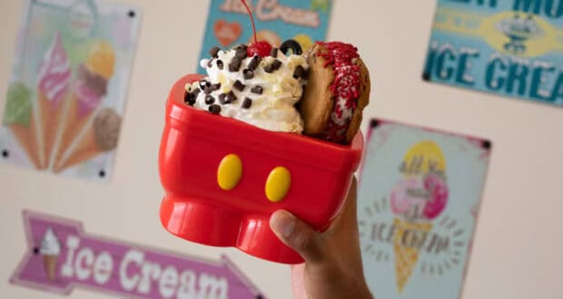 A hand holds a red cartoon-themed bowl brimming with ice cream, adorned with whipped cream, chocolate chips, cherries, and cookies—reminiscent of the whimsical Disney World ice cream treats. In the background, colorful ice cream posters decorate the wall.