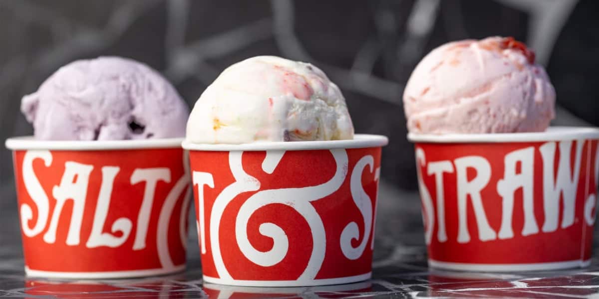 Three colorful scoops of ice cream are in branded red cups with "Salt & Straw" written on them, reminiscent of delightful Disney World ice cream treats. The scoops, in light purple, white, and pink flavors, stand out against a dark background with a subtle pattern.