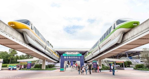 Disney World monorails glide above guests entering Magic Kingdom, passing under the iconic "Magic Kingdom Monorail" sign.