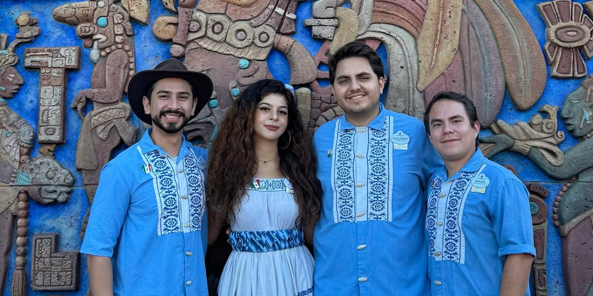Four people stand smiling in front of a vibrant, detailed mural with Aztec-style patterns. Three men, dressed as Disney World Cast Members in matching blue embroidered shirts, and a woman in a white dress with blue accents pose together.