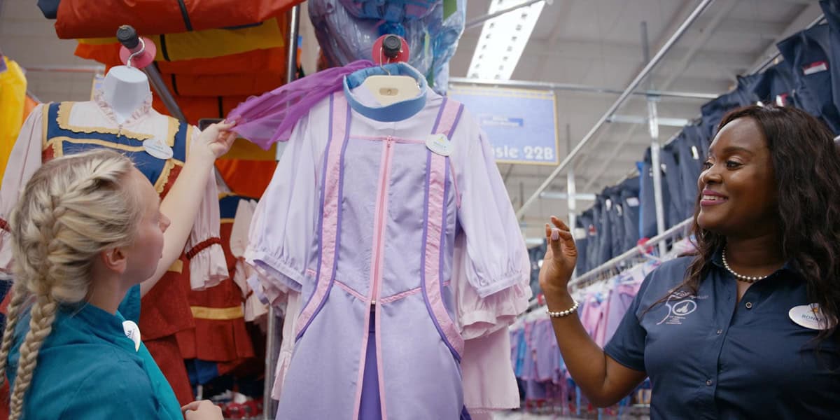 Two women, dressed as Disney World Cast Members, look at a mannequin in a light purple costume with pink trim. One lifts sheer purple fabric while the other smiles, both standing beside racks of clothing.