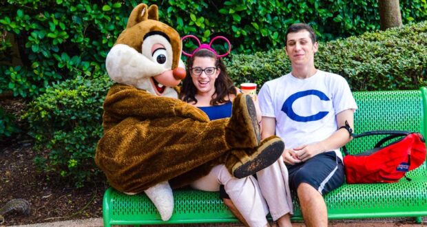 Two people sit on a green bench; one wears pink mouse ears and glasses, smiling next to a person in a chipmunk costume. The other person wears a white shirt with a blue "C." Shrubs and trees are in the background.