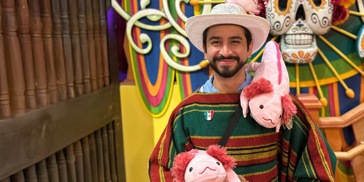A smiling man in a colorful poncho and white hat, reminiscent of Disney World Cast Members, wears two plush axolotls around his neck. He stands in front of festive, vibrant decorations, including a large sugar skull.