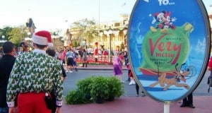 Festive guest poses by Mickey's Very Merry Christmas Party sign at Disney World entrance, kicking off 2025 holiday celebrations.
