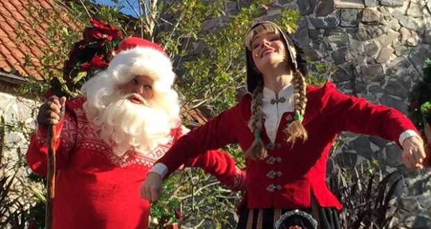 Santa and a Disney Cast Member in holiday costumes pose and smile by a stone building, celebrating festive magic at Disney Parks.