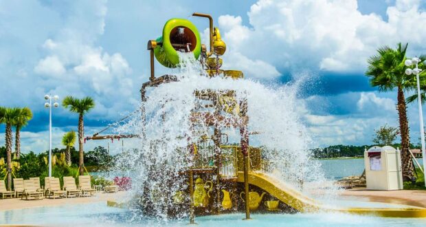 A large water play structure with slides and a giant bucket dumping water in a splash zone, surrounded by palm trees, lounge chairs, and a scenic lake under a partly cloudy sky.