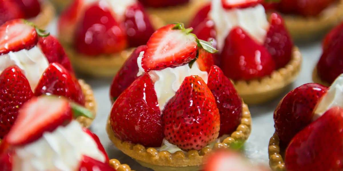 Close-up of mini tarts topped with fresh strawberries and dollops of whipped cream, reminiscent of the elegant treats found at EPCOT counter-service restaurants. Each tart is surrounded by tiny golden pearls, enhancing the vibrant red, glossy strawberries.