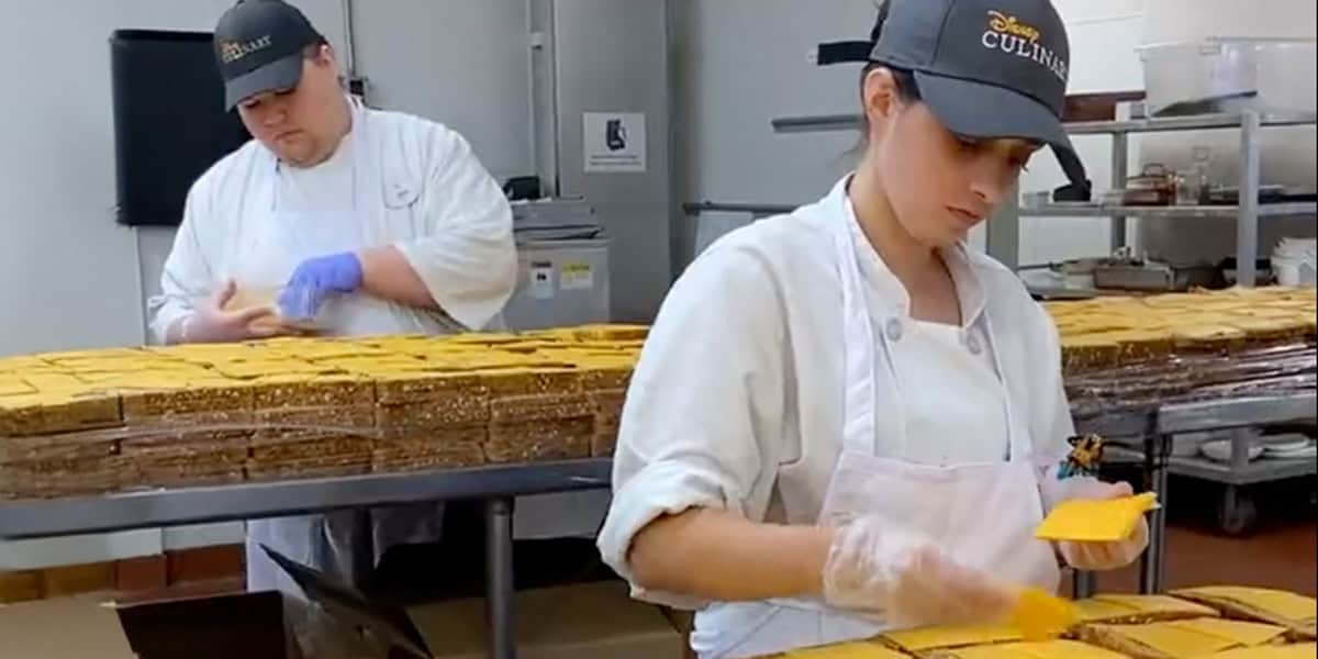 Two Disney World Cast Members in white uniforms and hats place slices of cheese on rows of sandwiches in an industrial kitchen with stainless steel shelves and equipment in the background.