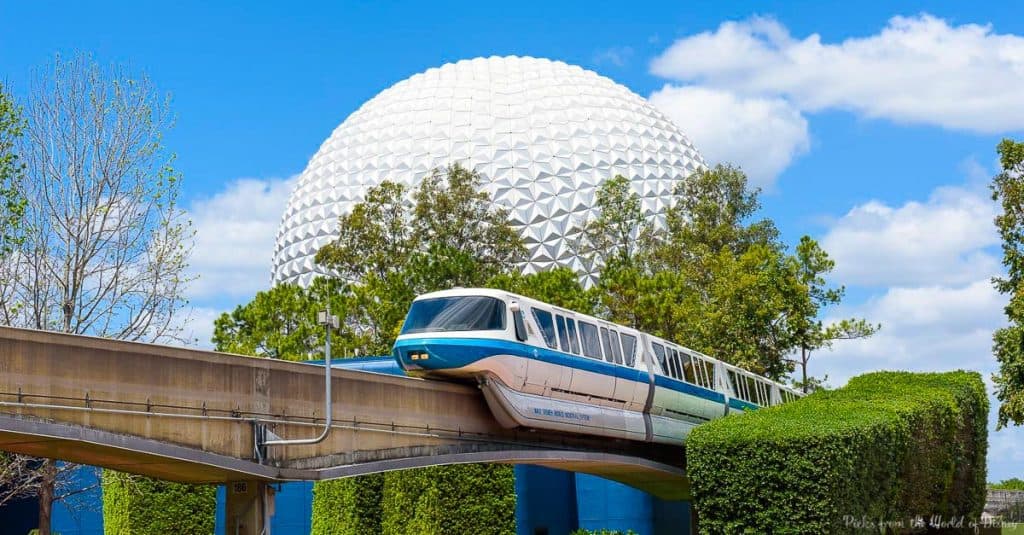 Disney’s iconic monorail glides past Spaceship Earth at Epcot, framed by lush trees and a brilliant blue sky.