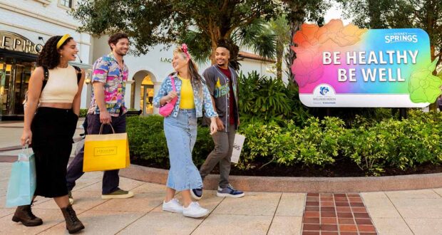 Four young adults walk outside, smiling and holding shopping bags, near a large colorful sign that says “Be Healthy Be Well.” They chat about Disney Shopping Tips as they stroll through the vibrant shopping area with stores and trees in the background.