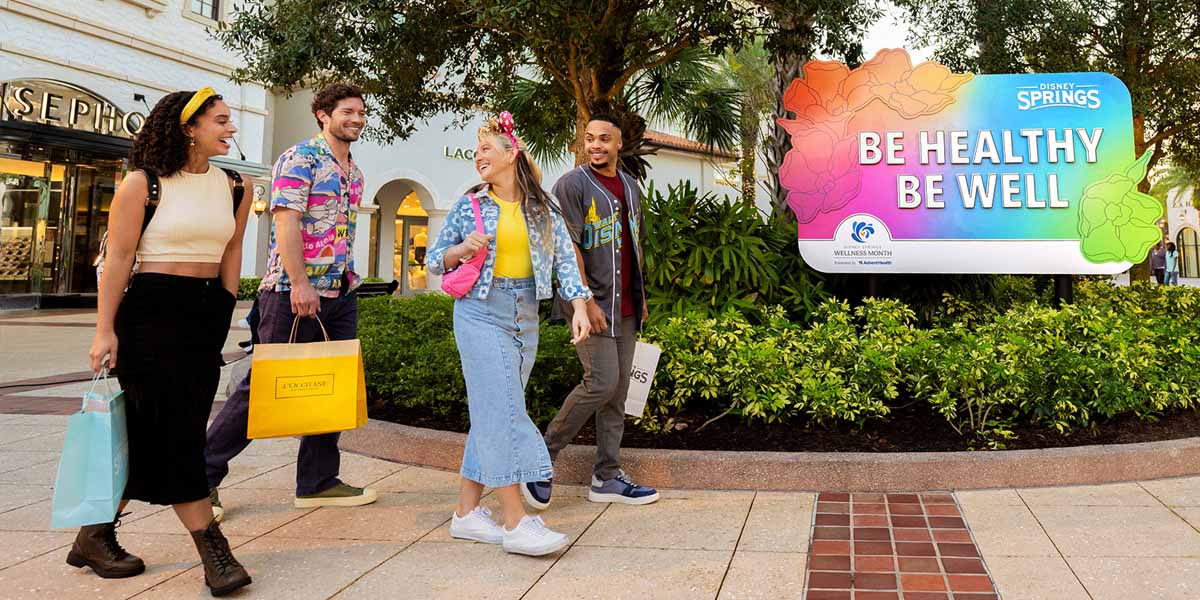 Four young adults walk outside, smiling and holding shopping bags, near a large colorful sign that says “Be Healthy Be Well.” They chat about Disney Shopping Tips as they stroll through the vibrant shopping area with stores and trees in the background.