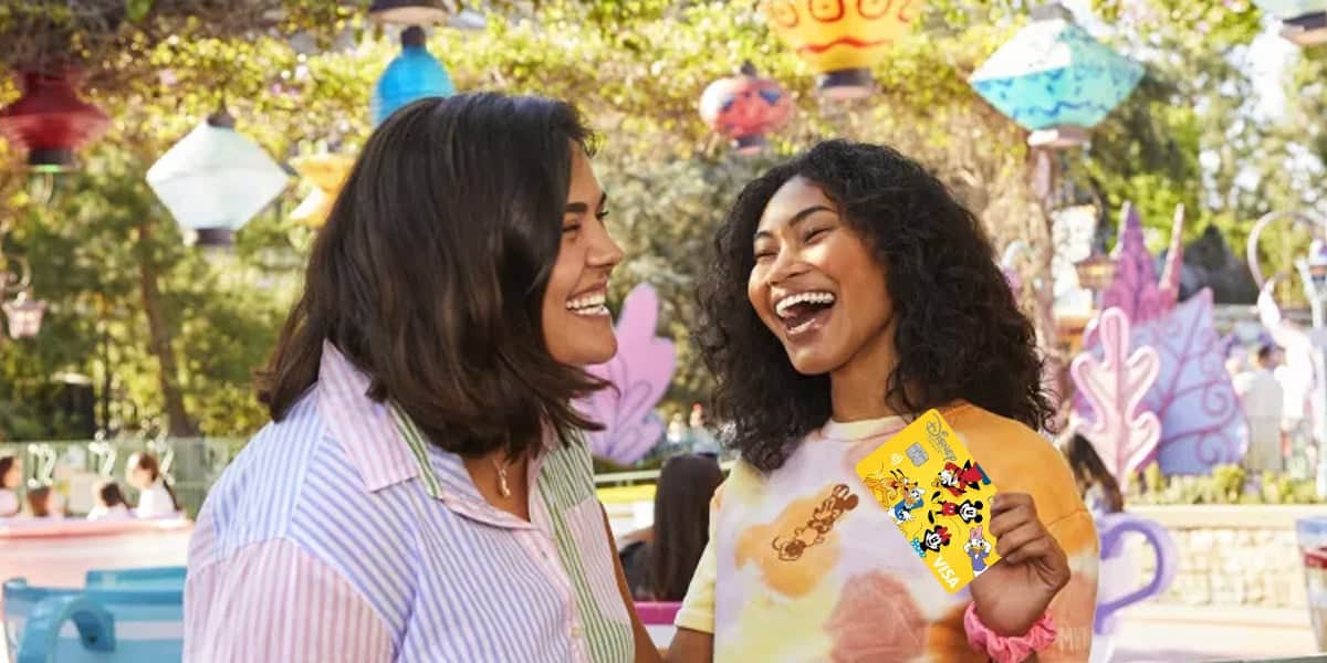 Two women smiling and laughing outdoors at an amusement park; one woman holds a yellow Disneyland ticket decorated with Disney characters. Colorful lanterns hang in the background, perfect for sharing Disney Shopping Tips.