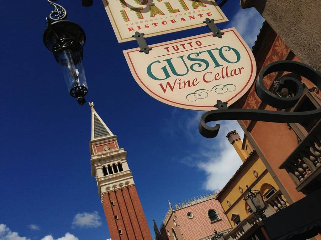 Tutto Gusto Wine Cellar sign and classic Italian facades at Epcot’s Italy Pavilion beneath a vibrant blue sky with fluffy clouds.