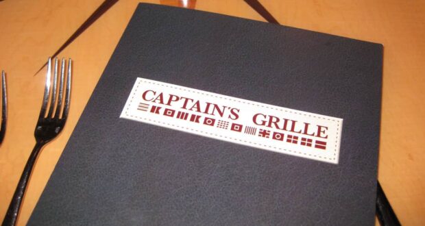 A dark menu cover labeled "Captain's Grille" with nautical signal flags, placed on a wooden table next to two forks.