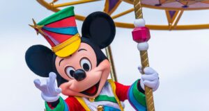 Mickey Mouse, in a colorful outfit and striped hat, waves cheerfully while standing on a parade float adorned with vibrant decorations. The background is a clear blue sky.