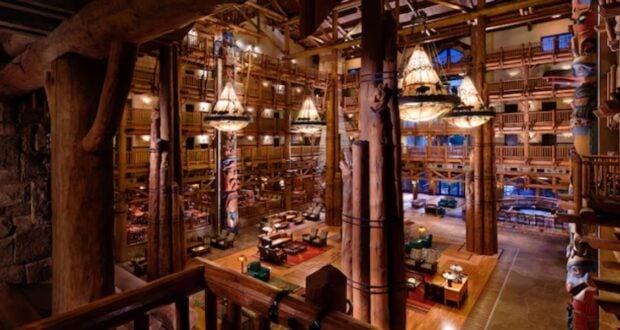 Spacious lodge interior with high ceilings, large wooden beams, and chandeliers. The room features wooden furniture, vibrant rugs, and ornate pillars. A mezzanine level overlooks the scene, adding to the rustic ambiance.