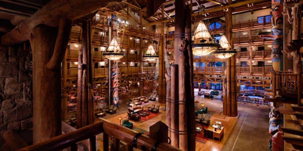 Spacious lodge interior with high ceilings, large wooden beams, and chandeliers. The room features wooden furniture, vibrant rugs, and ornate pillars. A mezzanine level overlooks the scene, adding to the rustic ambiance.