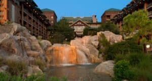 A rustic lodge with a triangular roof stands in the background, surrounded by lush greenery. In the foreground, a waterfall cascades over large rocks into a serene pool, creating a picturesque scene.