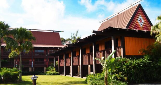 A tropical resort features buildings with steep, pointed roofs and wooden accents, surrounded by lush green plants and palm trees under a bright blue sky.