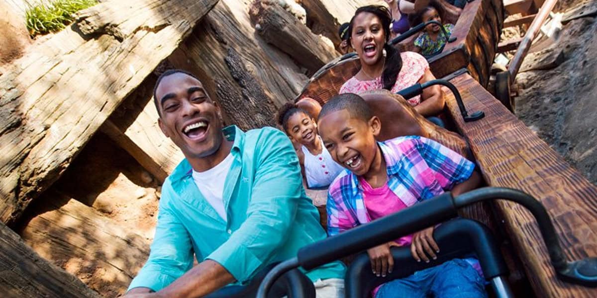 A family of four, smiling and laughing, rides a wooden roller coaster together on a sunny day. With Walt Disney World tips in mind, they make the most of the thrill, the father and son in front and mother and daughter right behind.