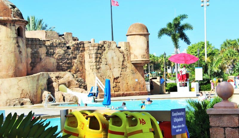 Caribbean Beach Resort pool with iconic stone castle, swaying palms, lifeguard, and yellow life jackets—relax Disney style!.