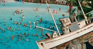 Lively pool scene with swimmers and a tilted pirate ship on rocks, evoking classic Disney water parks and adventure-themed resorts.