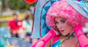 A vibrant Cast Member in a bright pink wig playfully shares secrets, delighting guests as crowds blur in the background.
