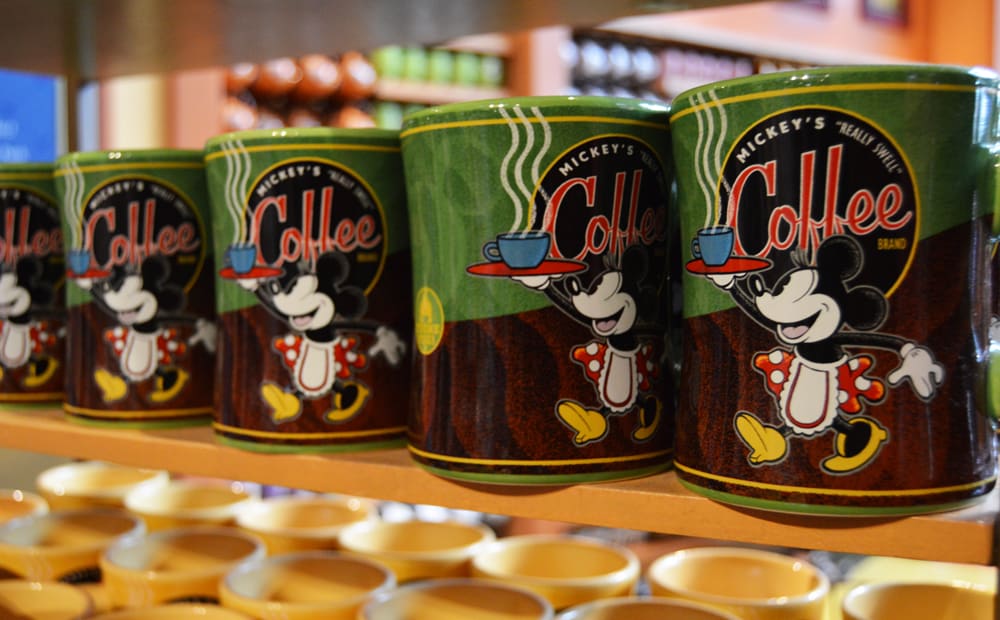 Mickey's Really Swell Coffee green mugs with Mickey Mouse are lined up on a Disney World shop shelf—perfect park souvenirs.