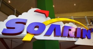 Soarin’ Around the World" sign with clouds and yellow arc, indoors by railing and green pillar—pure Disney Parks adventure vibes!.