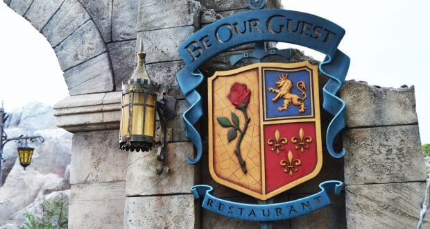 The iconic Be Our Guest Restaurant sign with crest glows by a lantern on a stone wall, inviting Disney fans to dine in style.