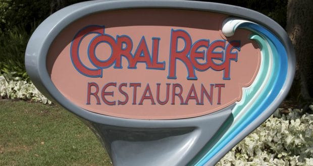 Coral Reef Restaurant sign with aquatic theme stands by vibrant gardens, welcoming Epcot guests for an underwater dining adventure.