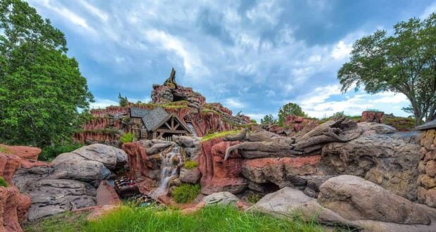 A log flume ride descends a steep drop on a rocky, red and brown mountain surrounded by greenery and trees under a partly cloudy sky.