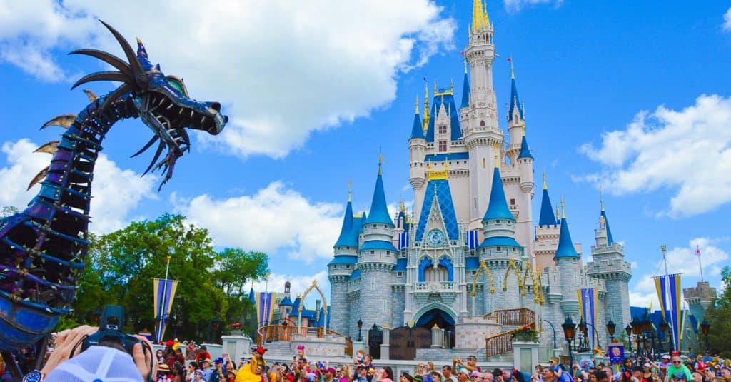 A large metal dragon float passes in front of Cinderella’s Castle at Disney World during a parade, with crowds of people gathered under a bright blue sky with white clouds.