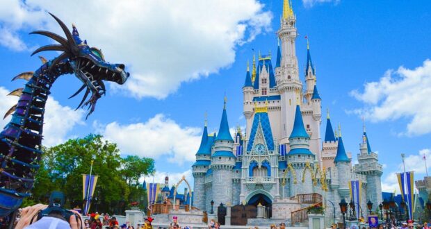 A large metal dragon float passes in front of Cinderella’s Castle at Disney World during a parade, with crowds of people gathered under a bright blue sky with white clouds.