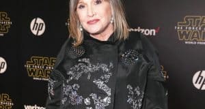 Carrie Fisher lookalike with gray hair in a black embroidered outfit at the Star Wars premiere, posing by film logos.