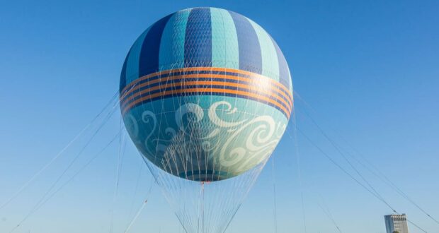 A large, colorful hot air balloon with blue and orange stripes is tethered to the ground with cables, floating above buildings under a clear blue sky.