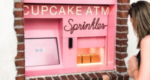 A woman stands in front of a pink Sprinkles cupcake ATM built into a brick wall. Three cupcake boxes are visible inside the machine’s open window.